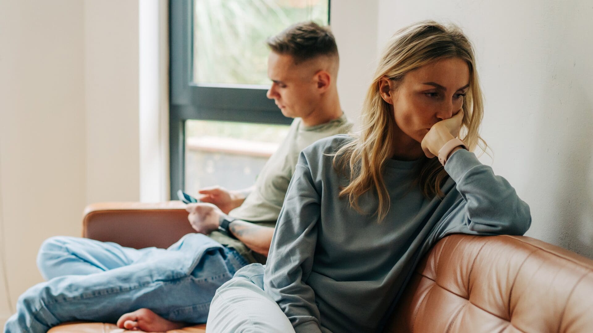 Woman sitting on a sofa with a worried expression, turned away from her partner who is also facing away, showing emotional distance and relationship tension.