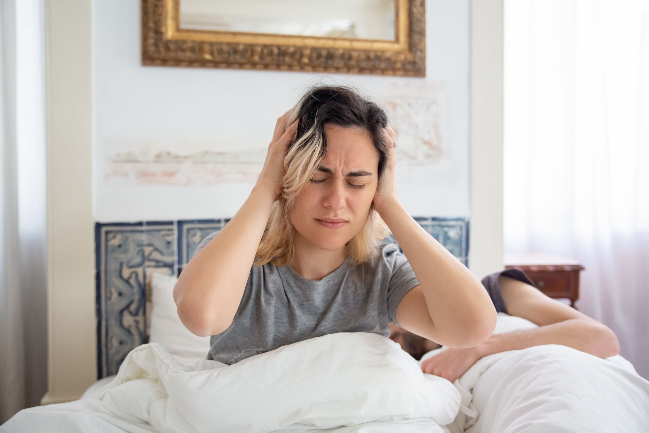 Woman awake sitting on bed with her head in her hands while her partner sleeps beside her.